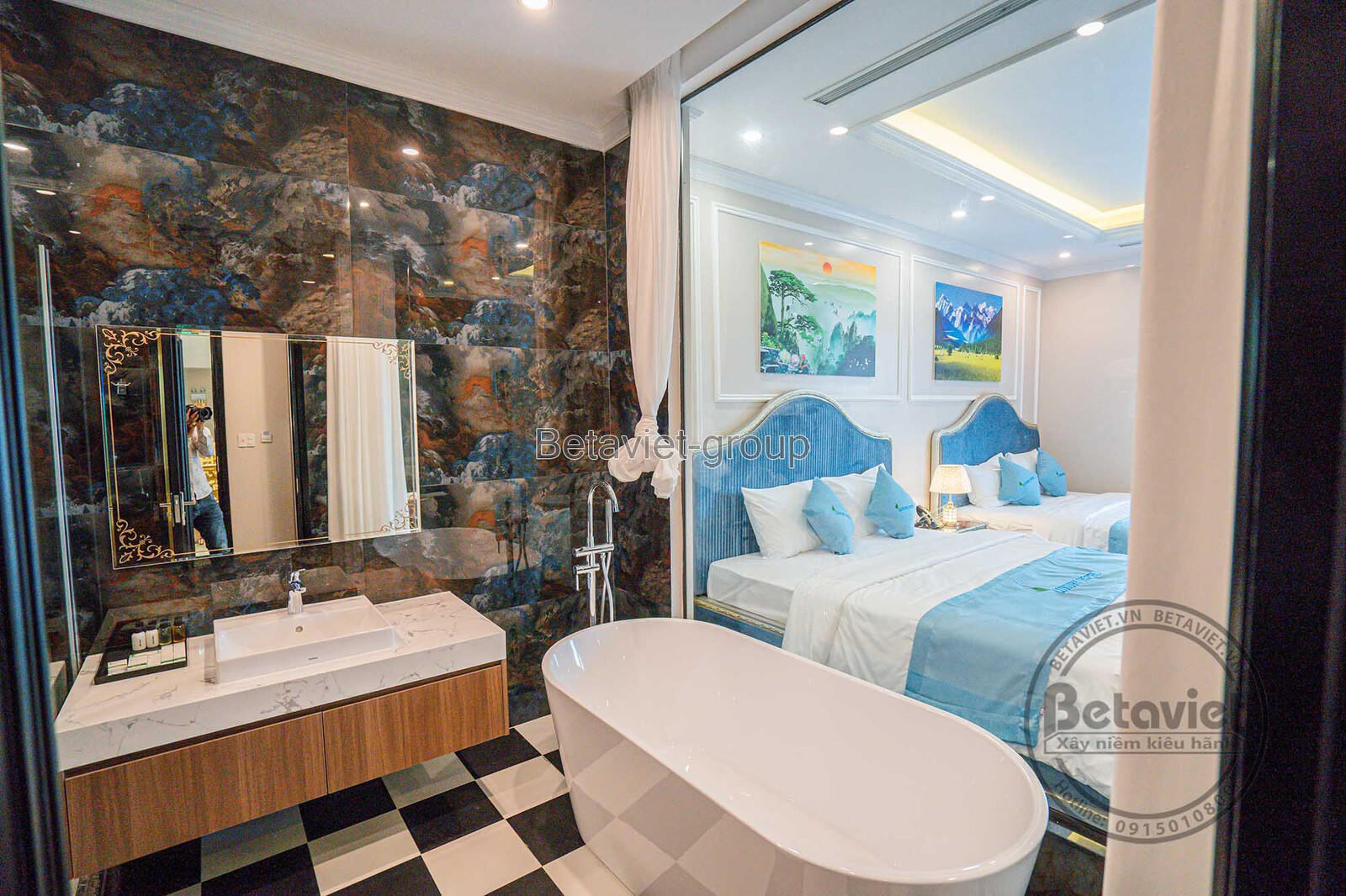 Khu vực bồn rửa mặt với gương lớn, tủ gỗ dưới bồn và tường ốp đá marble đen vân vàng. Bồn tắm độc lập màu trắng đặt cạnh…