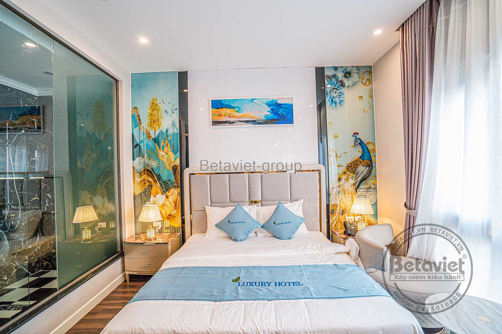 Phòng ngủ khách sạn với giường đôi bọc nệm đầu giường màu kem, ga trải giường trắng xanh. Tranh treo tường phong cảnh và đ…