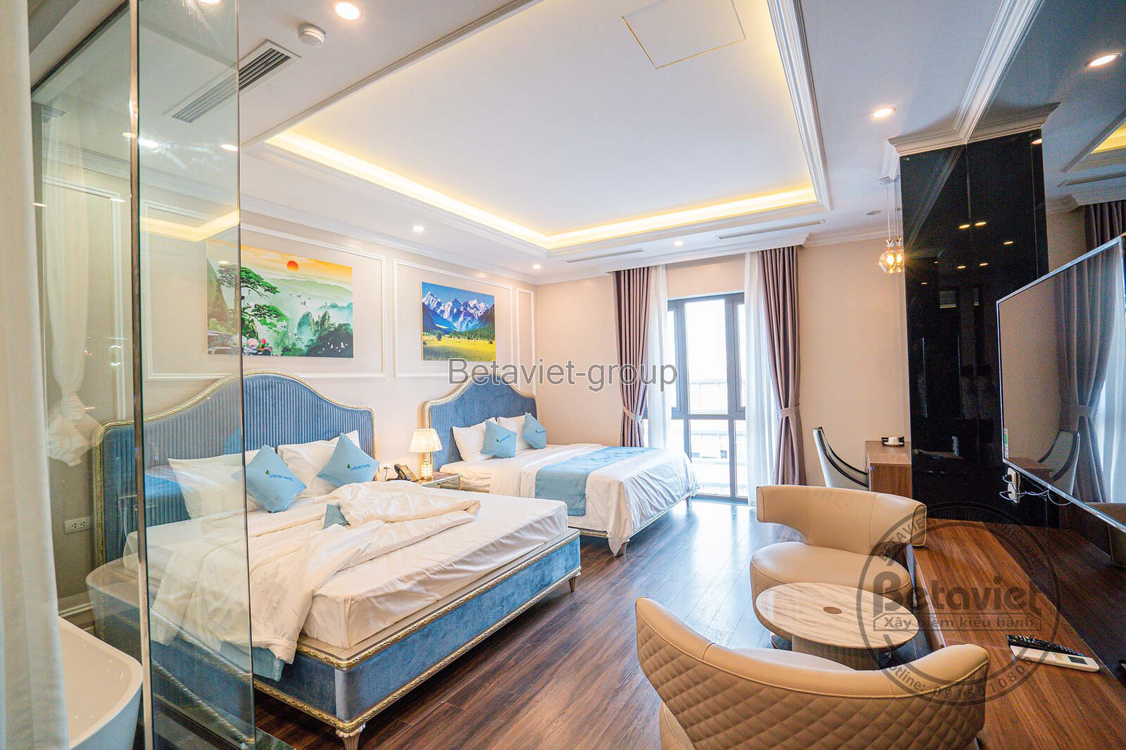 Phòng ngủ khách sạn rộng rãi với giường đôi lớn, sofa dài màu kem và kệ TV treo tường. Trần thạch cao giật cấp với đèn hắt…