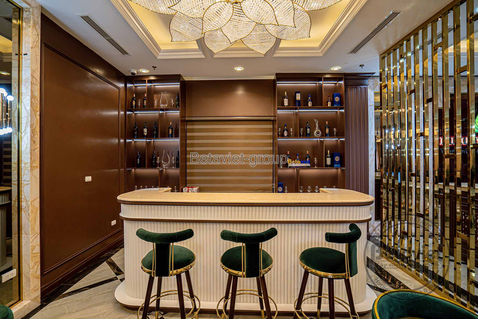 Khu vực quầy bar mini với tủ rượu gỗ màu nâu sẫm, kệ trưng bày chai rượu và ly cốc. Ghế bar bọc nhung xanh đậm và ánh sáng…
