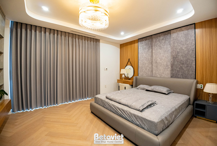 Phòng ngủ với giường lớn bọc nỉ màu xám, tủ đầu giường trắng. Tường ốp gỗ và nỉ, sàn gỗ xương cá. Ánh sáng ấm áp, tạo khôn…