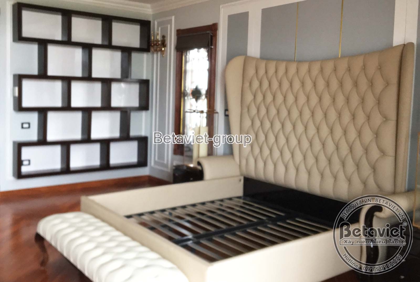 Không gian phòng ngủ đang lắp đặt nội thất gỗ tối màu, với vách đầu giường chạm khắc tinh xảo và tủ kệ trang trí, tạo điểm…