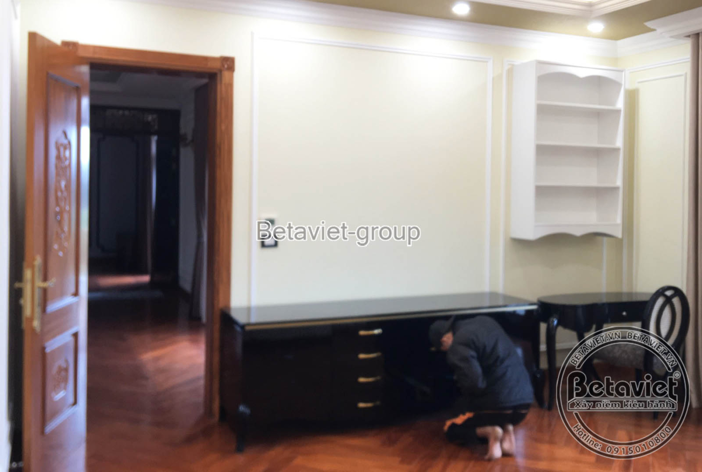 Không gian phòng ngủ đang lắp đặt nội thất gỗ tối màu, với giường bọc nệm màu kem và tủ đầu giường, tạo sự hài hòa và tiện…