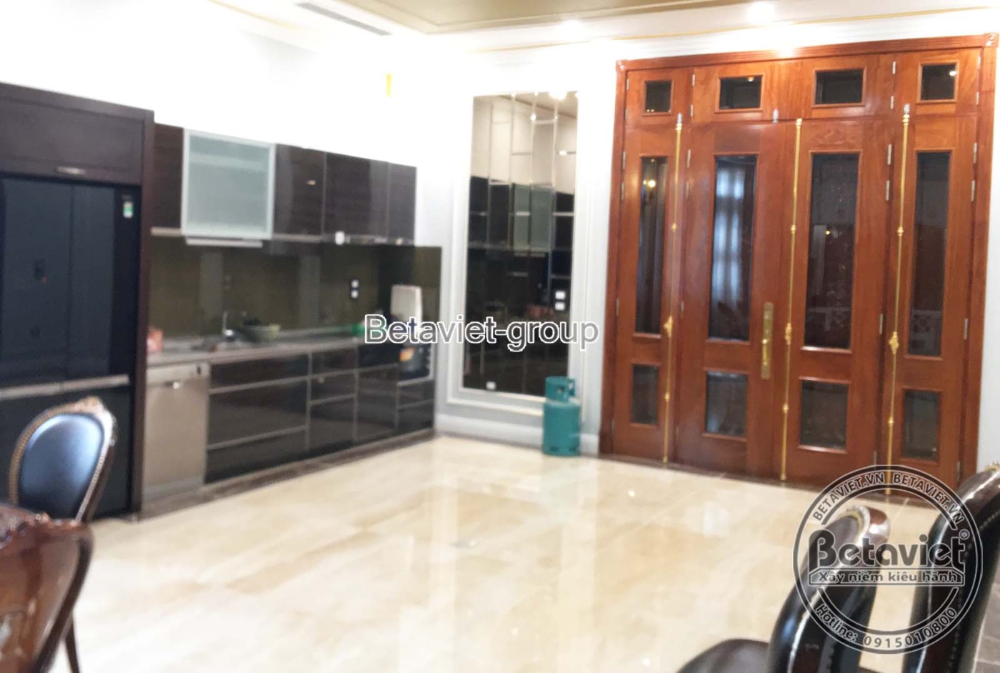 Không gian bếp hoặc phòng ăn đang lắp đặt tủ bếp và cửa gỗ lớn, với sàn gạch sáng màu, tạo không gian hiện đại và tiện ngh…