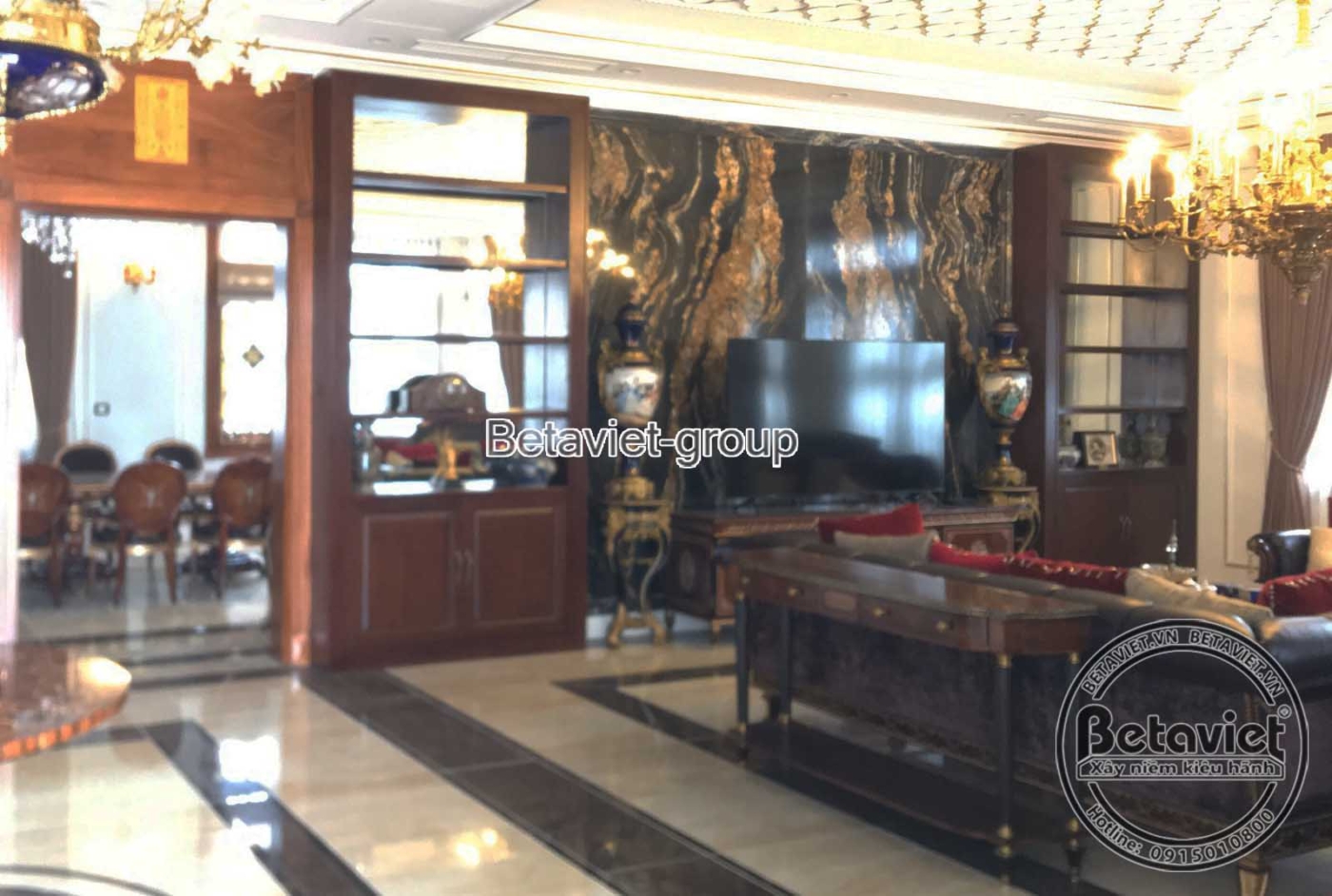 Không gian phòng khách hoặc phòng sinh hoạt chung với nội thất gỗ tối màu, sàn đá hoa văn và đèn chùm pha lê, mang phong c…