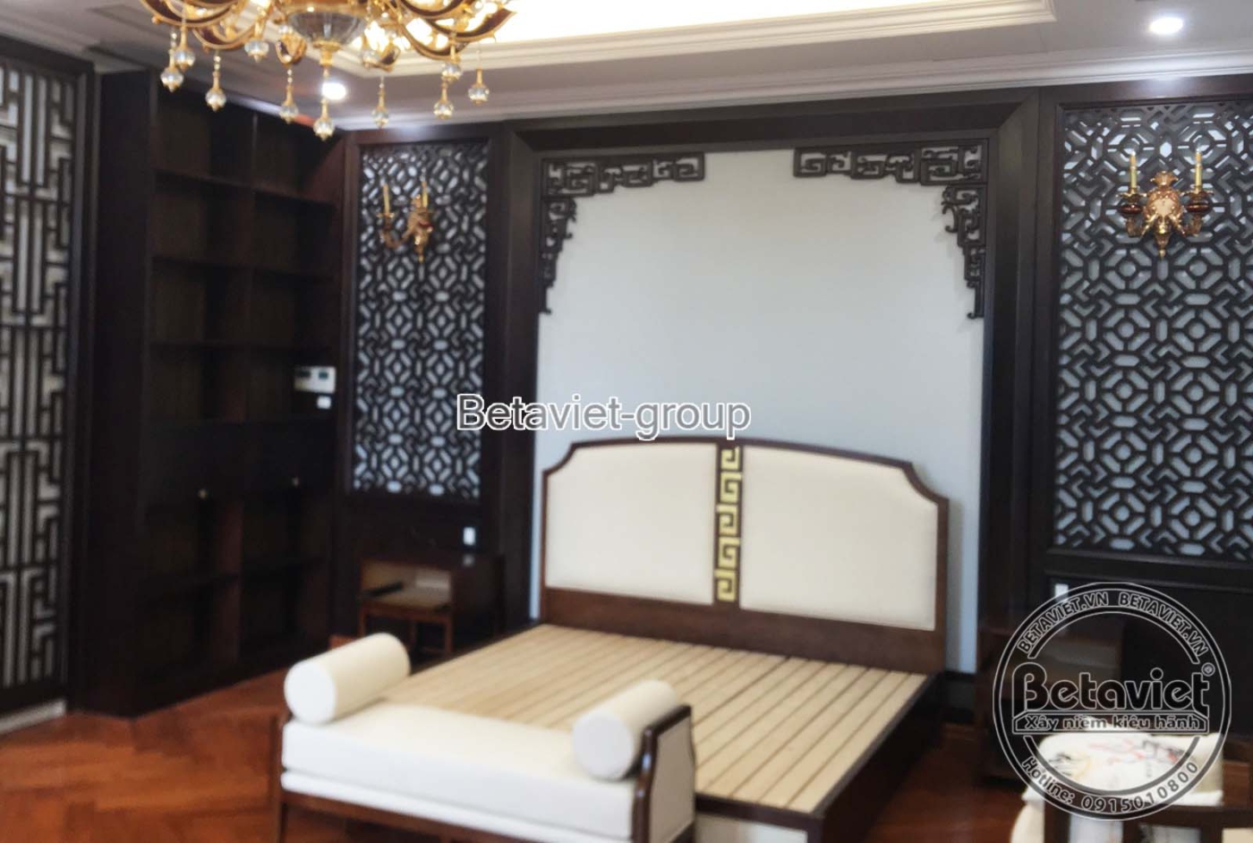Không gian phòng ngủ đang lắp đặt nội thất gỗ tối màu, với vách đầu giường chạm khắc và tủ kệ trang trí, tạo điểm nhấn Á Đ…