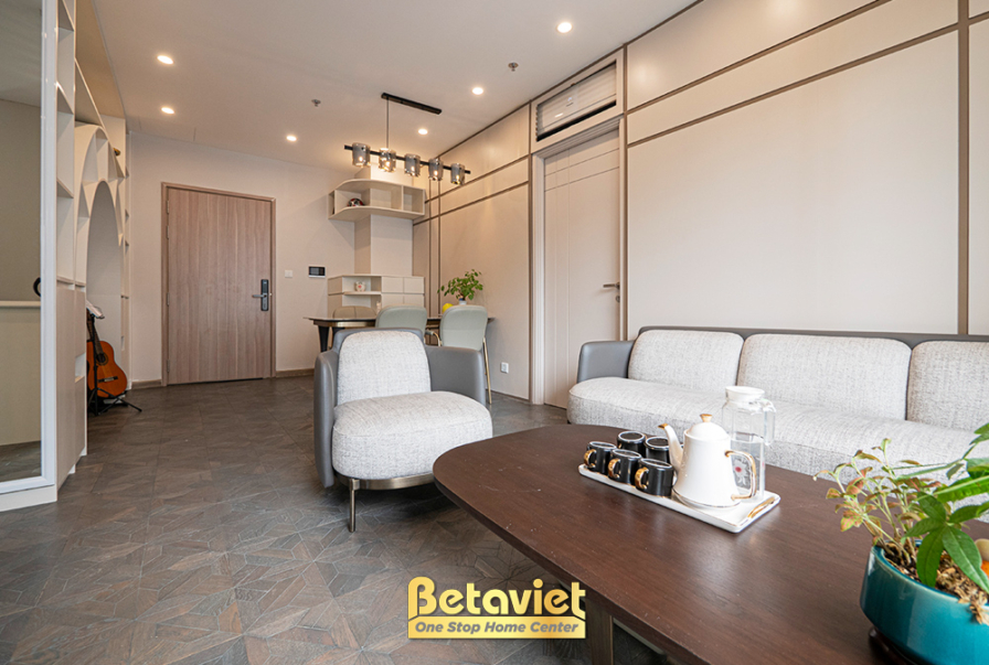 Toàn cảnh phòng khách và khu vực lối vào căn hộ. Sofa nỉ màu be kết hợp bàn trà gỗ. Tường ốp phào chỉ dọc màu be và kệ tra…