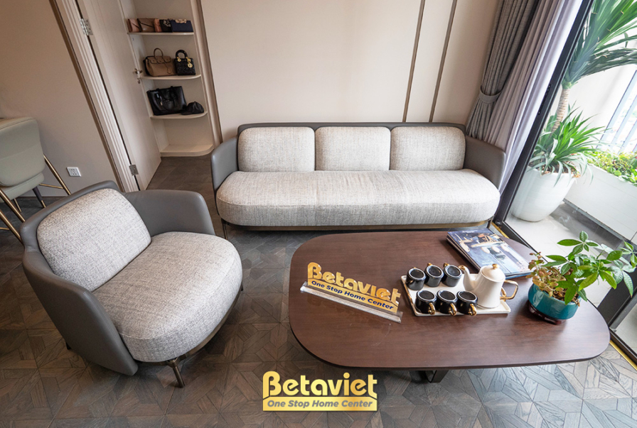 Cận cảnh khu vực sofa trong phòng khách. Sofa nỉ màu ghi nhạt với gối tựa màu xanh và be. Bàn trà gỗ mặt đá được trang trí…
