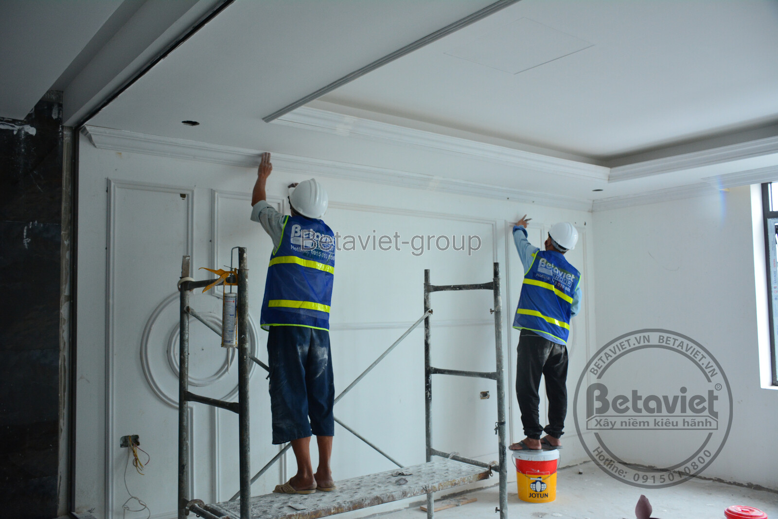 Hai công nhân đang thi công lắp đặt phào chỉ trần và tường, chuẩn bị cho việc sơn bả và hoàn thiện chi tiết kiến trúc. Khô…