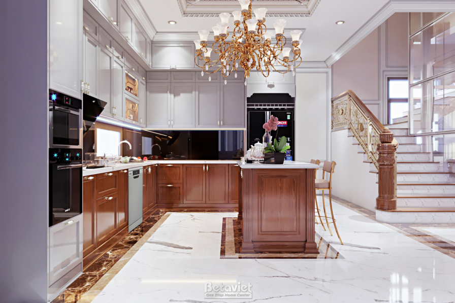 Bếp tủ gỗ nâu, thiết bị âm tủ như lò nướng, đảo bếp mặt đá marble, cầu thang tay vịn đồng phía sau, không gian sáng thoáng.