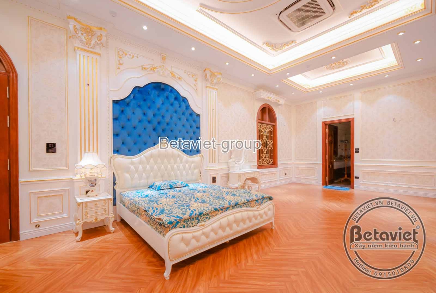 Phòng ngủ master phong cách tân cổ điển với giường lớn bọc nệm, đầu giường ốp gỗ chạm khắc và nệm màu xanh ngọc. Tường ốp…