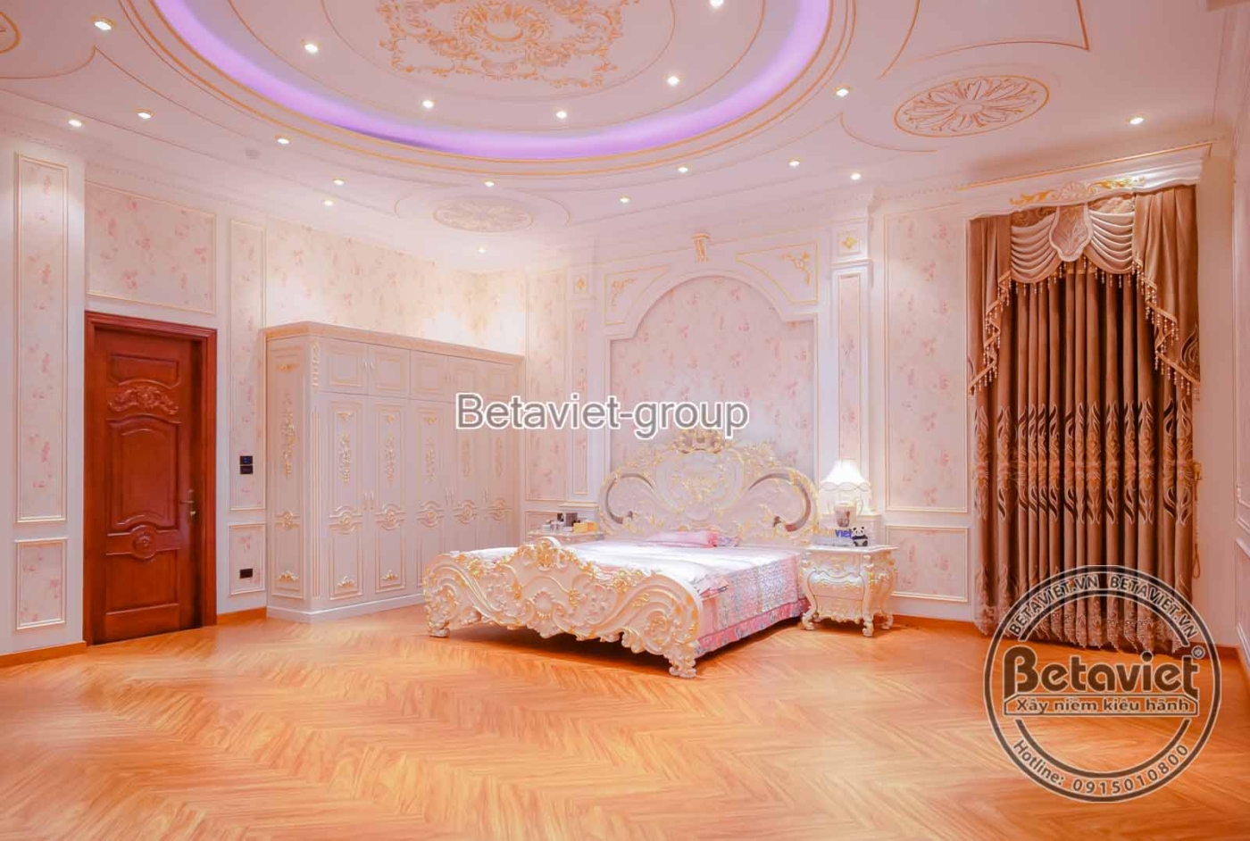 Phòng ngủ với giường lớn bọc nệm trắng, đầu giường chạm khắc tinh xảo. Tường ốp phào chỉ và giấy dán tường họa tiết nhẹ nh…