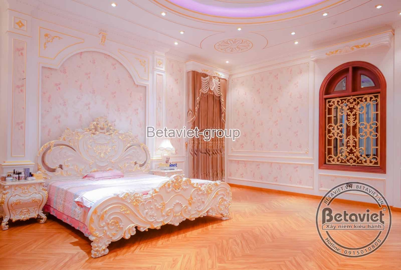 Phòng ngủ với giường lớn, đầu giường bọc nệm và chạm khắc tinh tế. Tường ốp phào chỉ và giấy dán tường họa tiết, cửa sổ vò…