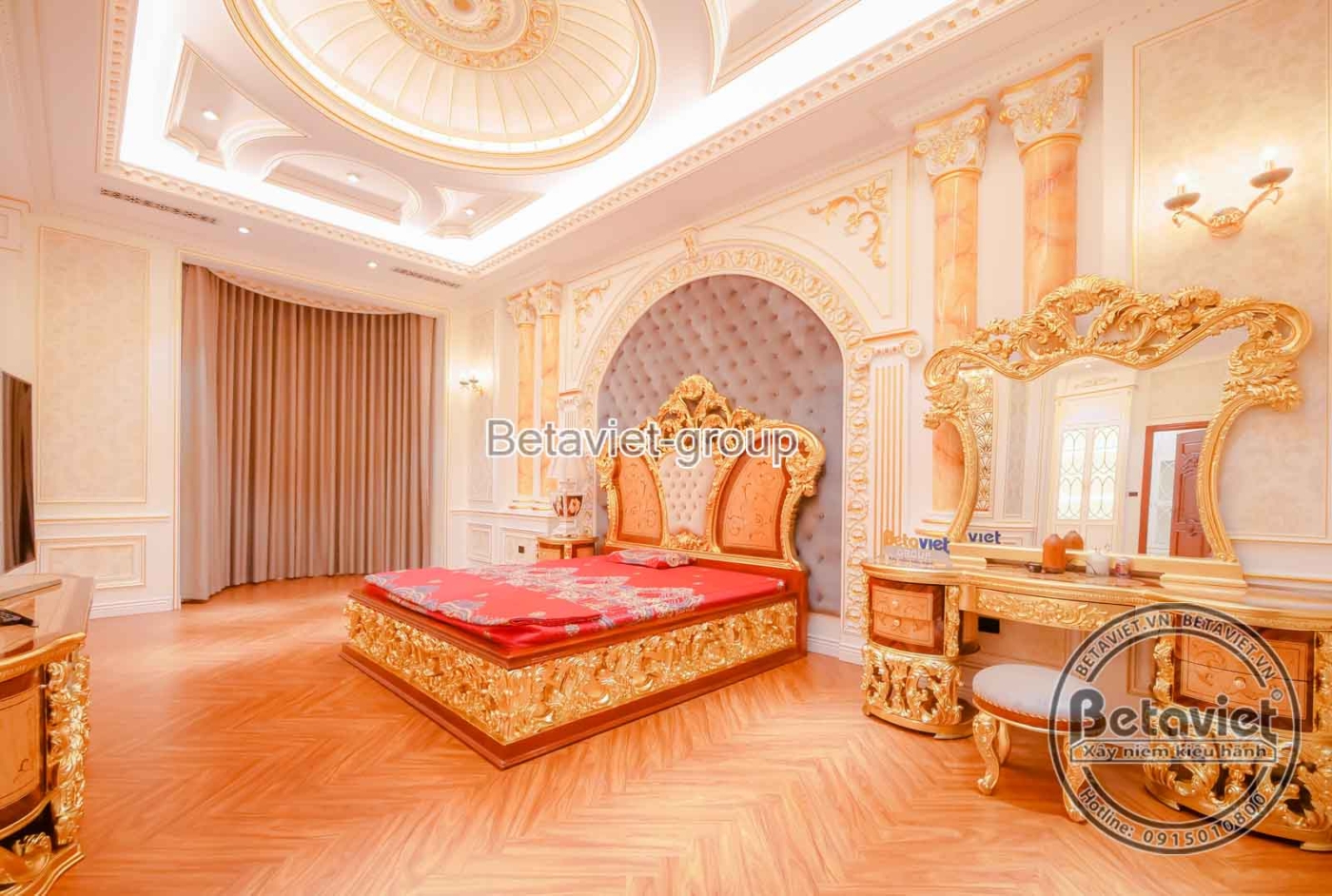 Phòng ngủ với giường lớn màu đỏ nổi bật, đầu giường bọc nệm và khung gỗ chạm khắc cầu kỳ. Bàn trang điểm cổ điển với gương…