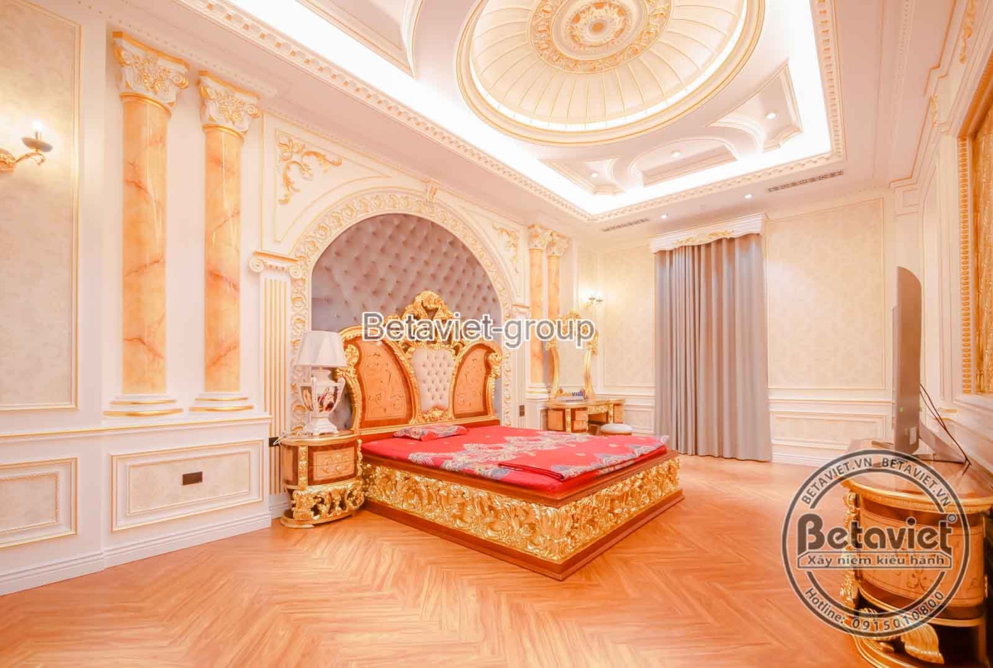 Góc nhìn khác của phòng ngủ màu đỏ, tập trung vào giường và bàn trang điểm. Ánh sáng tự nhiên từ cửa sổ lớn kết hợp với án…