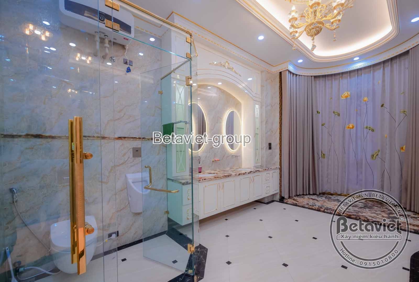 Khu vực lavabo trong phòng tắm, với tủ lavabo gỗ màu trắng kem, mặt đá sáng bóng. Gương lớn viền cổ điển, vách kính tắm đứ…