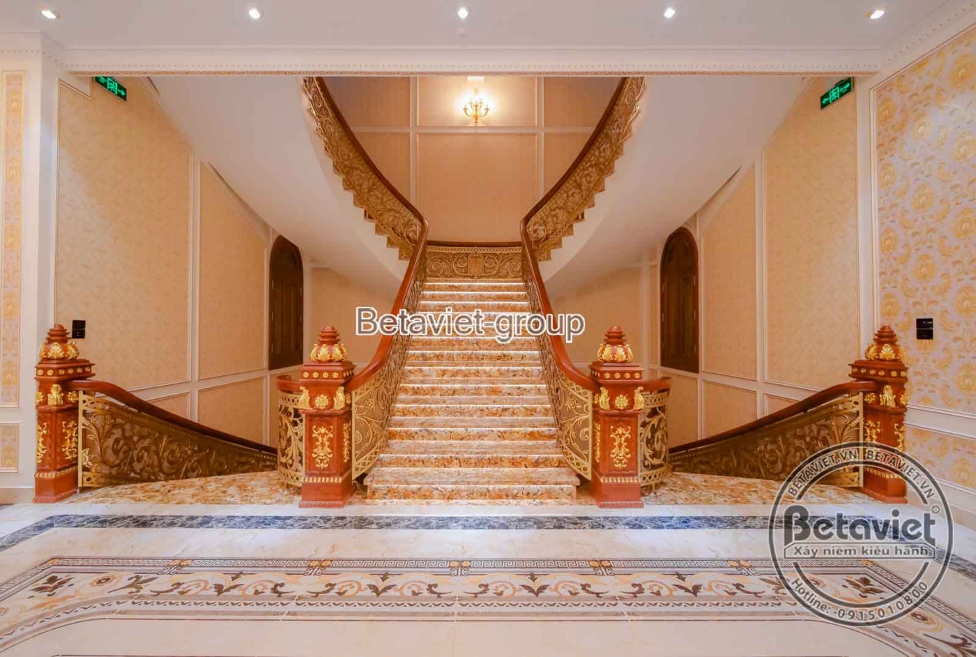Cầu thang chính với lan can gỗ và con tiện đá, bậc thang lát đá vân mây sáng bóng. Tường ốp hoa văn dát vàng tinh xảo, trầ…