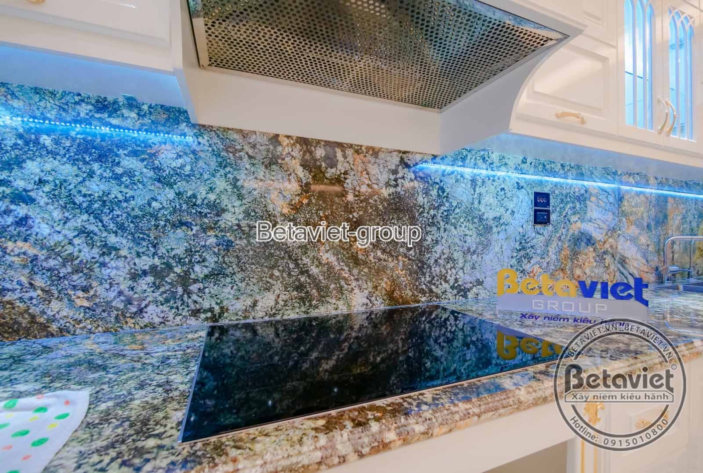 Khu vực bếp với tủ bếp dưới ốp đá granite màu xanh đen, mặt bếp và tường ốp đá vân sáng. Hệ thống hút mùi hiện đại phía tr…