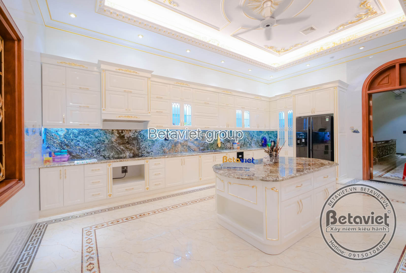 Tủ bếp trên màu trắng kem với phào chỉ cổ điển, kết hợp mặt bếp và tường ốp đá vân. Bố cục gọn gàng, tối ưu công năng, ánh…