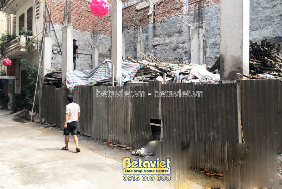 Công trường nhà phố nhìn từ đường phố, với các công nhân đang làm việc và vật liệu xây dựng chất đống. Xung quanh là các n…