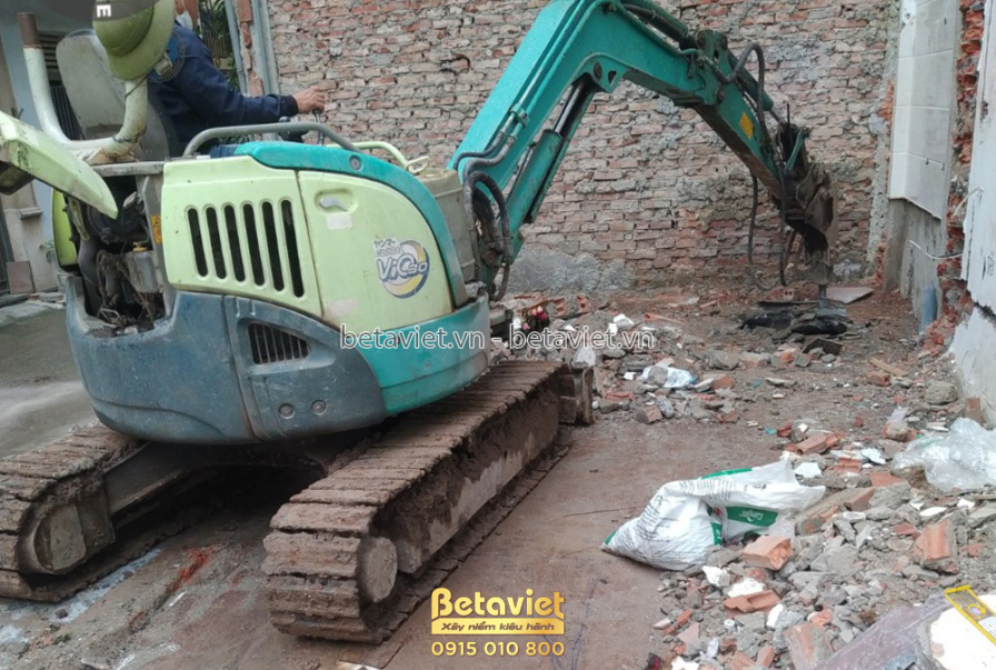Máy xúc đang hoạt động trên nền đất đỏ, xung quanh là gạch vỡ và đất đá. Đây là giai đoạn đào móng hoặc dọn dẹp mặt bằng…