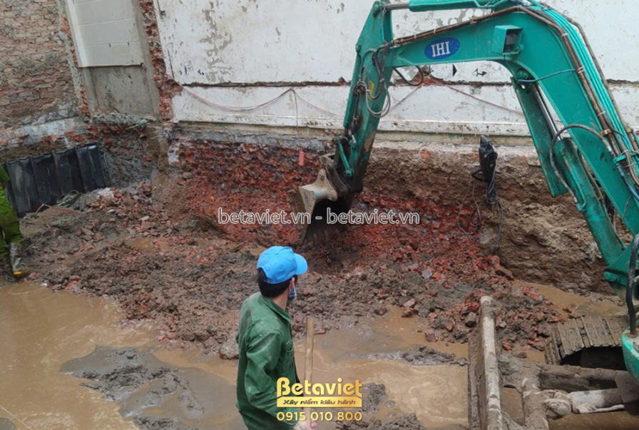 Công nhân đang đứng cạnh hố móng sâu, nền đất ẩm ướt và có nước đọng. Máy xúc đang tiếp tục đào đất, đảm bảo độ sâu và kíc…