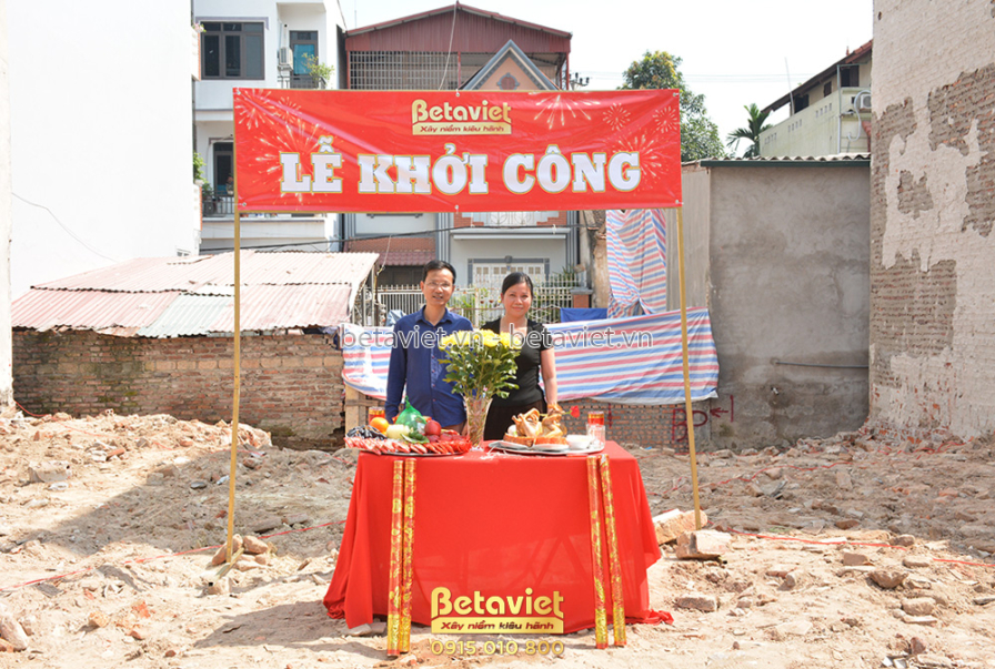 Lễ khởi công dự án với bàn cúng đặt giữa công trường, có hoa quả và đồ lễ. Phía sau là phông bạt đỏ lớn ghi "LỄ KHỞI CÔNG"…