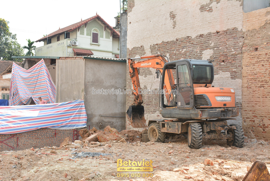 Cảnh công trường đang san lấp mặt bằng, một máy xúc màu cam đang hoạt động trên nền đất trống. Xung quanh là tường gạch cũ…