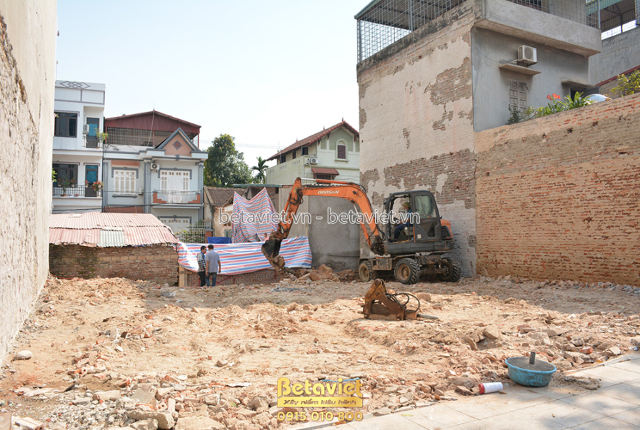 Toàn cảnh công trường đang được san lấp, máy xúc nhỏ đang làm việc. Xung quanh là các công trình lân cận và tường gạch. Đấ…
