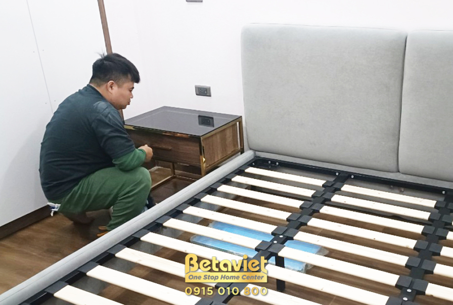 Công nhân đang lắp đặt nội thất phòng ngủ, cụ thể là phần giường ngủ bọc nệm và tủ đầu giường gỗ. Không gian phòng ngủ hiệ…