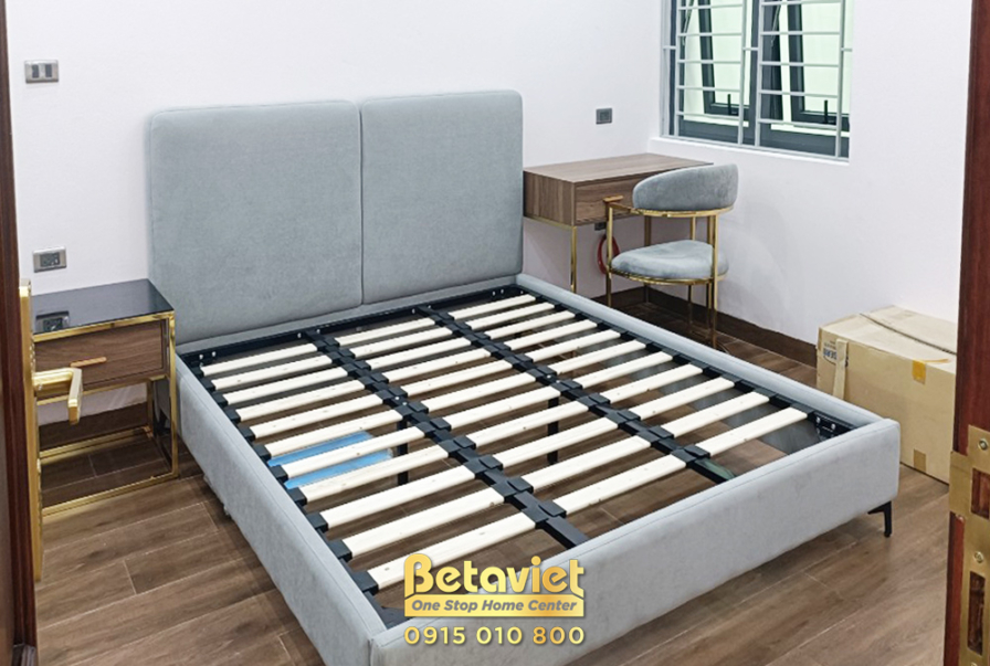 Phòng ngủ với giường bọc nệm màu xám, tủ đầu giường gỗ và bàn làm việc nhỏ. Cửa sổ lớn mang lại ánh sáng tự nhiên, làm sán…