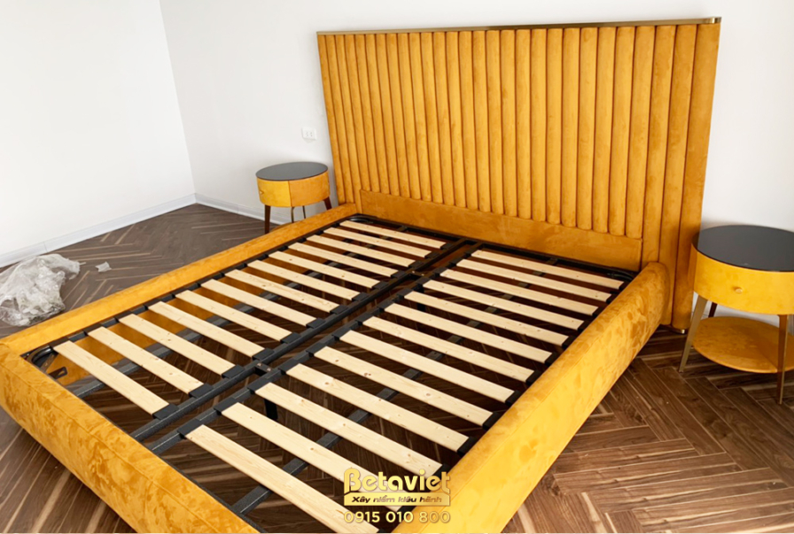 Giường ngủ đang lắp đặt với khung và đầu giường bọc nỉ màu vàng mustard nổi bật. Đầu giường thiết kế dạng panel dọc, kết h…
