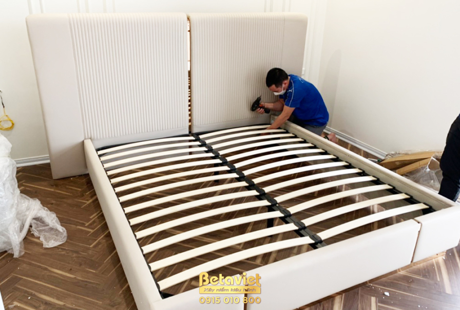 Công nhân đang hoàn thiện lắp đặt phần khung giường bọc nỉ màu be. Đầu giường cao, bọc nỉ với các đường may dọc, hứa hẹn m…