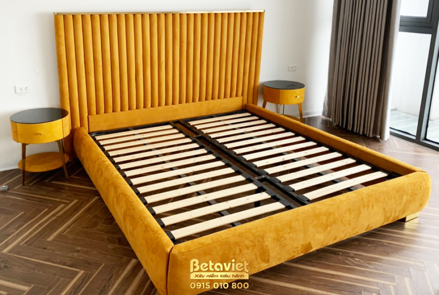 Giường ngủ đã lắp đặt hoàn thiện với khung và đầu giường bọc nỉ màu vàng mustard rực rỡ. Tab đầu giường tròn nhỏ màu vàng…