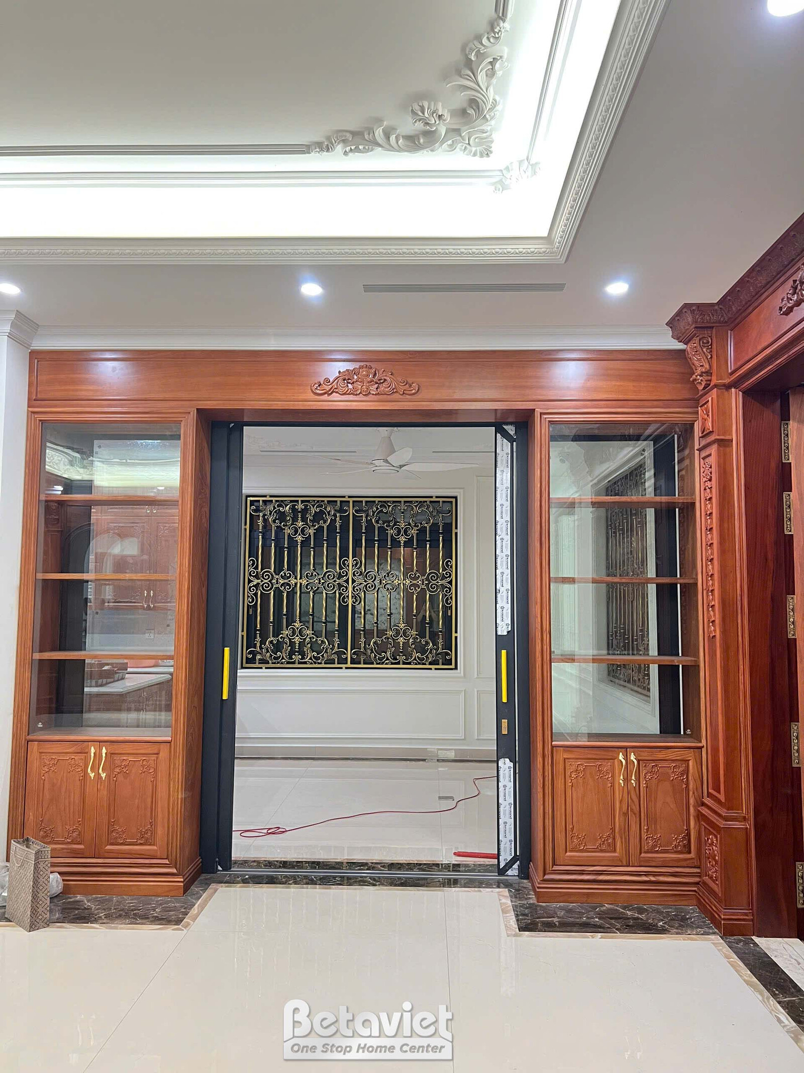 Lối vào phòng với cửa kính khung sắt hoa văn cổ điển, hai bên là tủ gỗ tự nhiên có kệ trưng bày đèn chiếu sáng. Trần thạch…