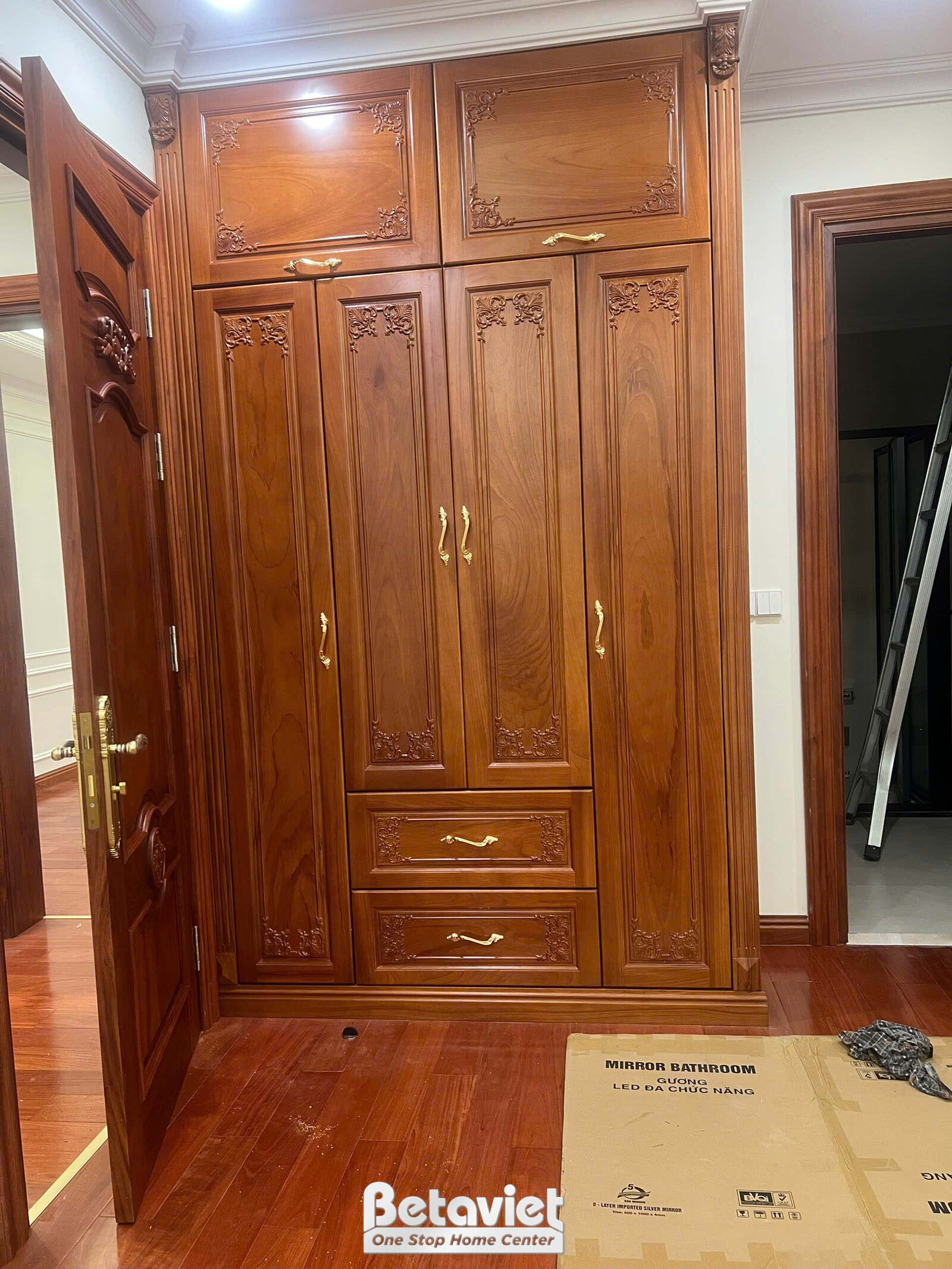 Sảnh cửa chính với bộ cửa gỗ tự nhiên nâu đỏ và tủ gương âm tường hai bên, tay nắm đồng ánh kim. Sàn gỗ nâu bóng, ánh sáng…