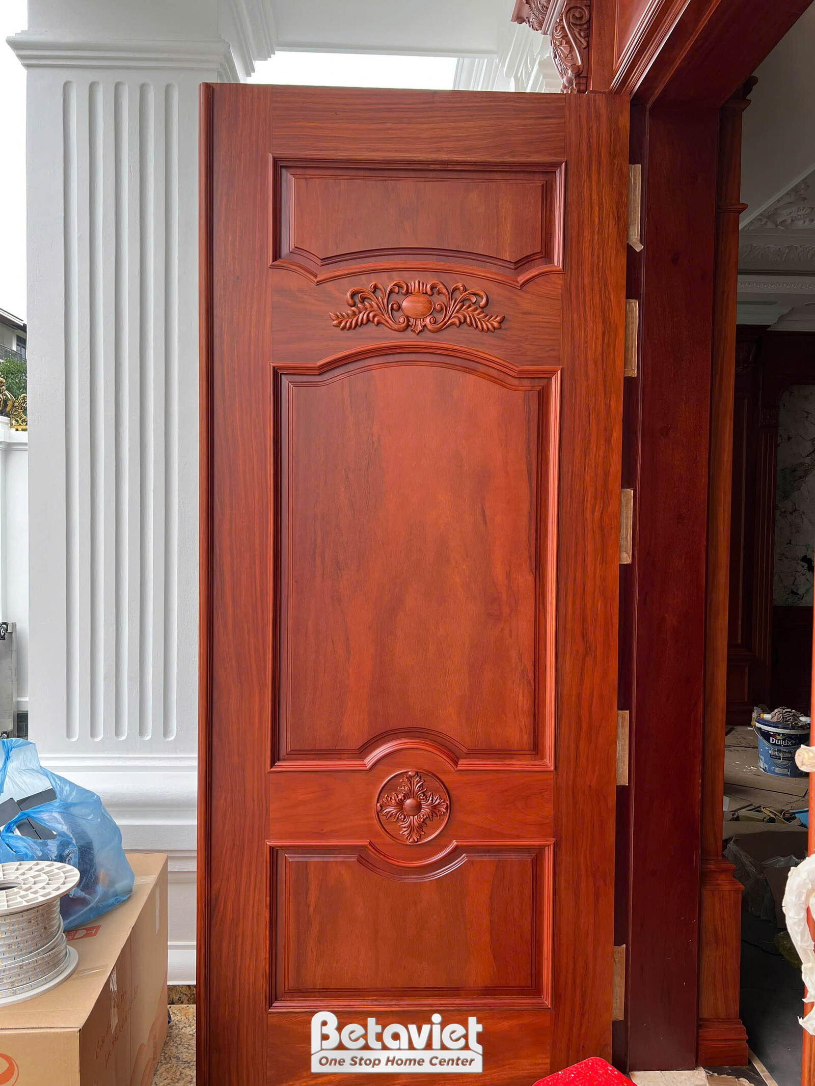 Cận cảnh cửa gỗ tự nhiên tân cổ điển chạm khắc hoa văn nổi trên cánh cửa màu nâu đỏ, khung bao gỗ và phào chỉ trắng ôm sát…