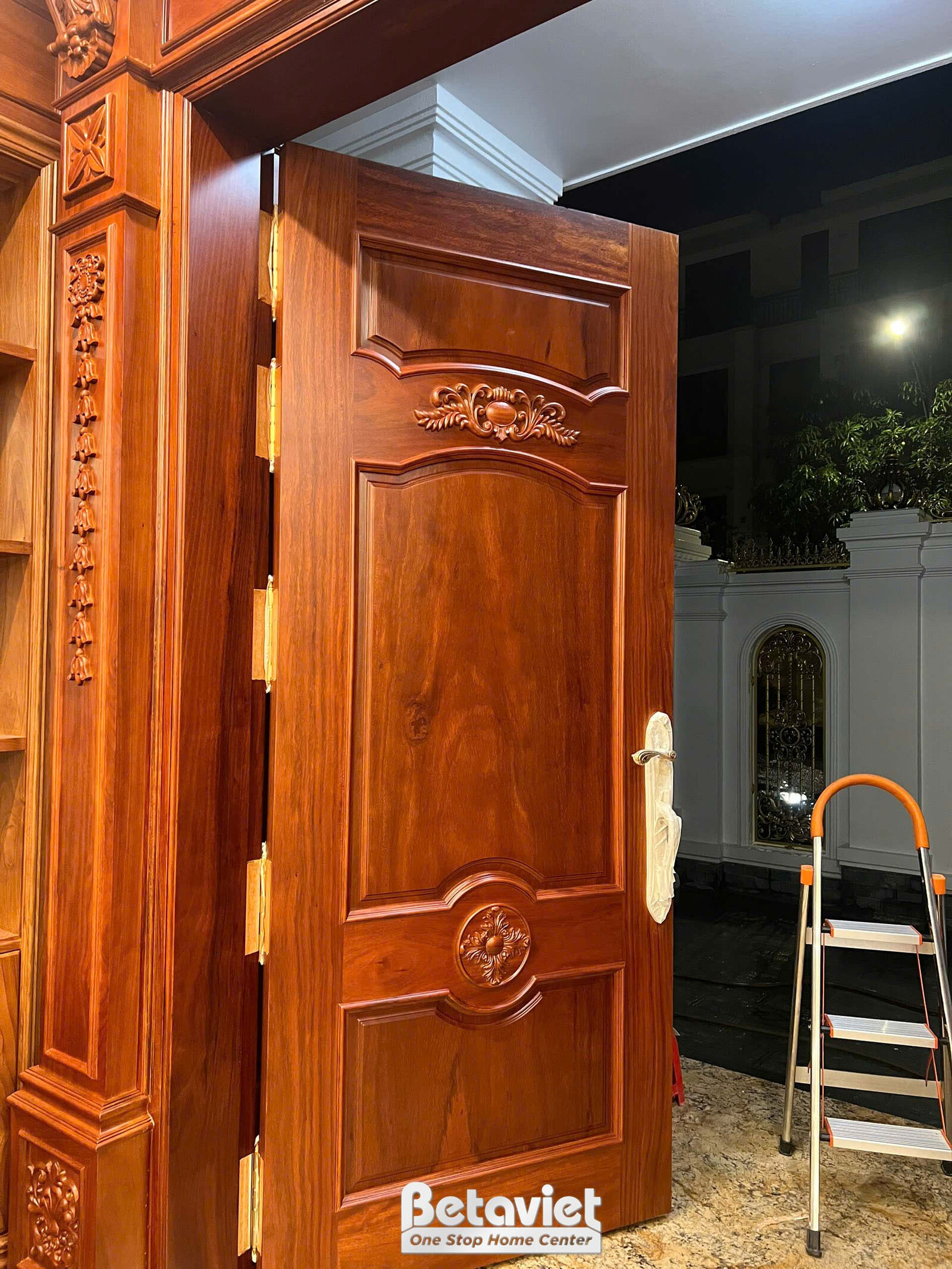 Cửa gỗ tự nhiên màu nâu đỏ với các họa tiết chạm khắc tinh xảo, đang trong quá trình lắp đặt hoàn thiện. Ánh sáng tự nhiên…