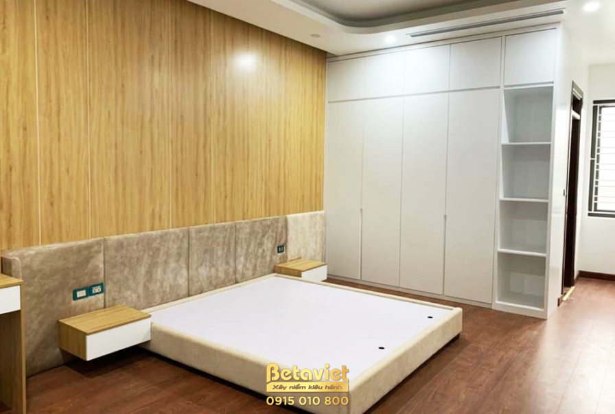 Phòng ngủ đã hoàn thiện với giường bục thấp màu trắng, vách đầu giường ốp gỗ màu vàng nhạt và vật liệu bọc nệm màu be. Tủ…