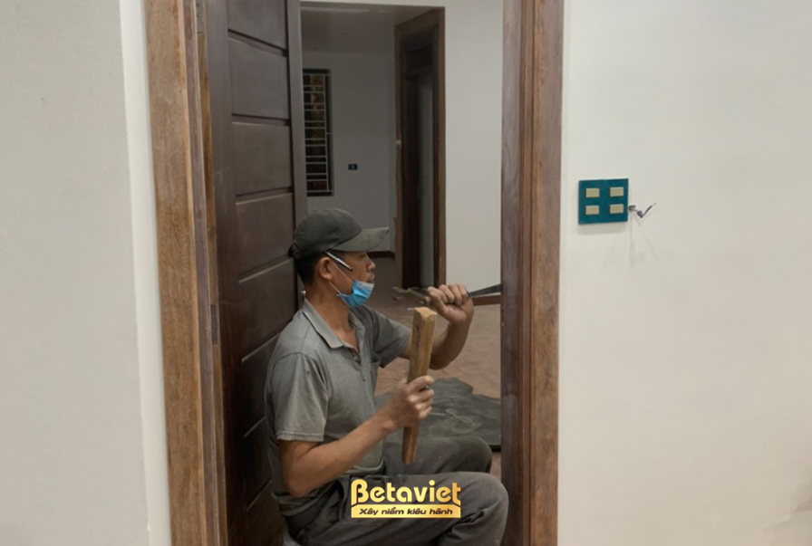 Cận cảnh công nhân đang tỉ mỉ lắp đặt khung cửa gỗ màu nâu trầm. Chi tiết công việc cho thấy sự cẩn trọng trong từng bước…