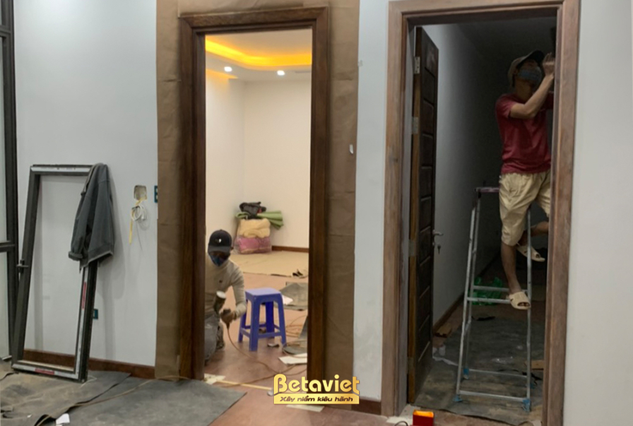 Hành lang hoặc khu vực cửa ra vào đang trong giai đoạn lắp đặt khung cửa gỗ. Sàn nhà đã được lát gạch sáng màu. Ánh sáng t…