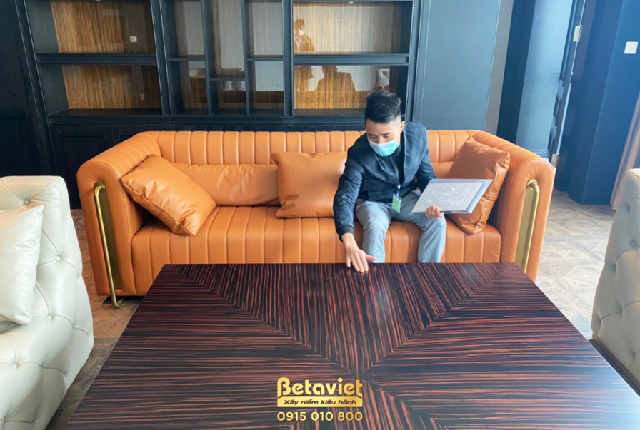 Góc phòng khách với sofa bọc nỉ màu cam đất, bàn trà lớn bằng gỗ tối màu. Bố cục ấm cúng, vật liệu nỉ và gỗ tạo cảm giác g…