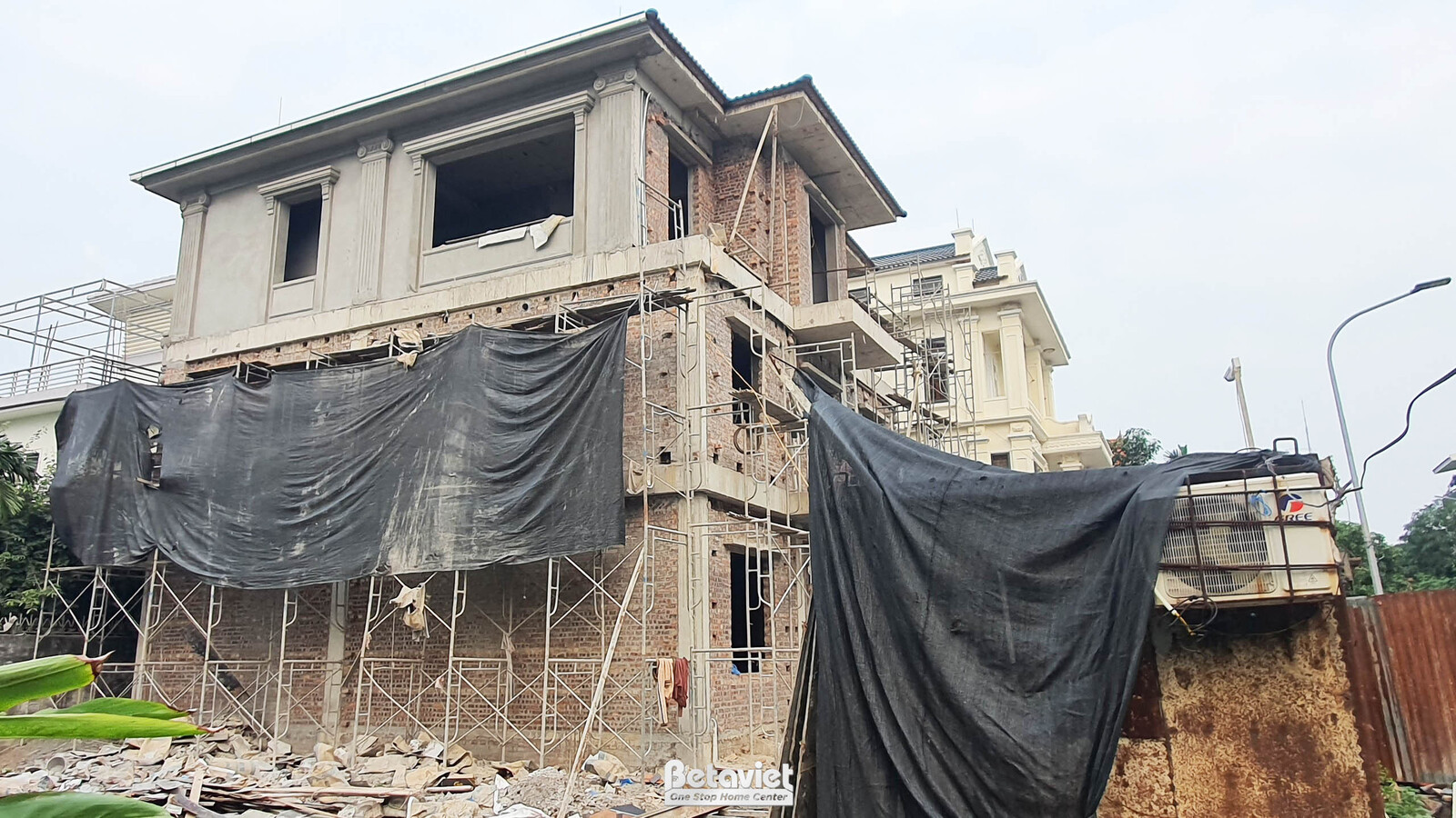 Ngôi nhà phố 3 tầng đang thi công phần thô, nhìn từ góc nghiêng. Lưới che chắn màu đen bao phủ một phần, giàn giáo vẫn còn…