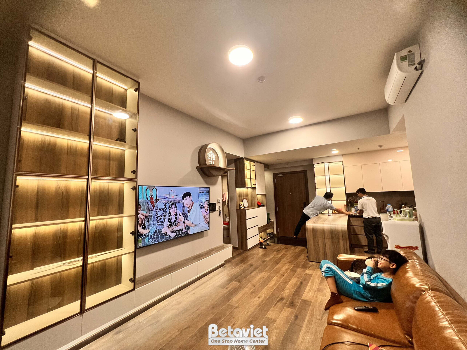 Góc nhìn tổng thể phòng khách đã hoàn thiện, với kệ tivi treo tường và sofa da màu nâu. Sàn gỗ tông ấm kết hợp nội thất mà…