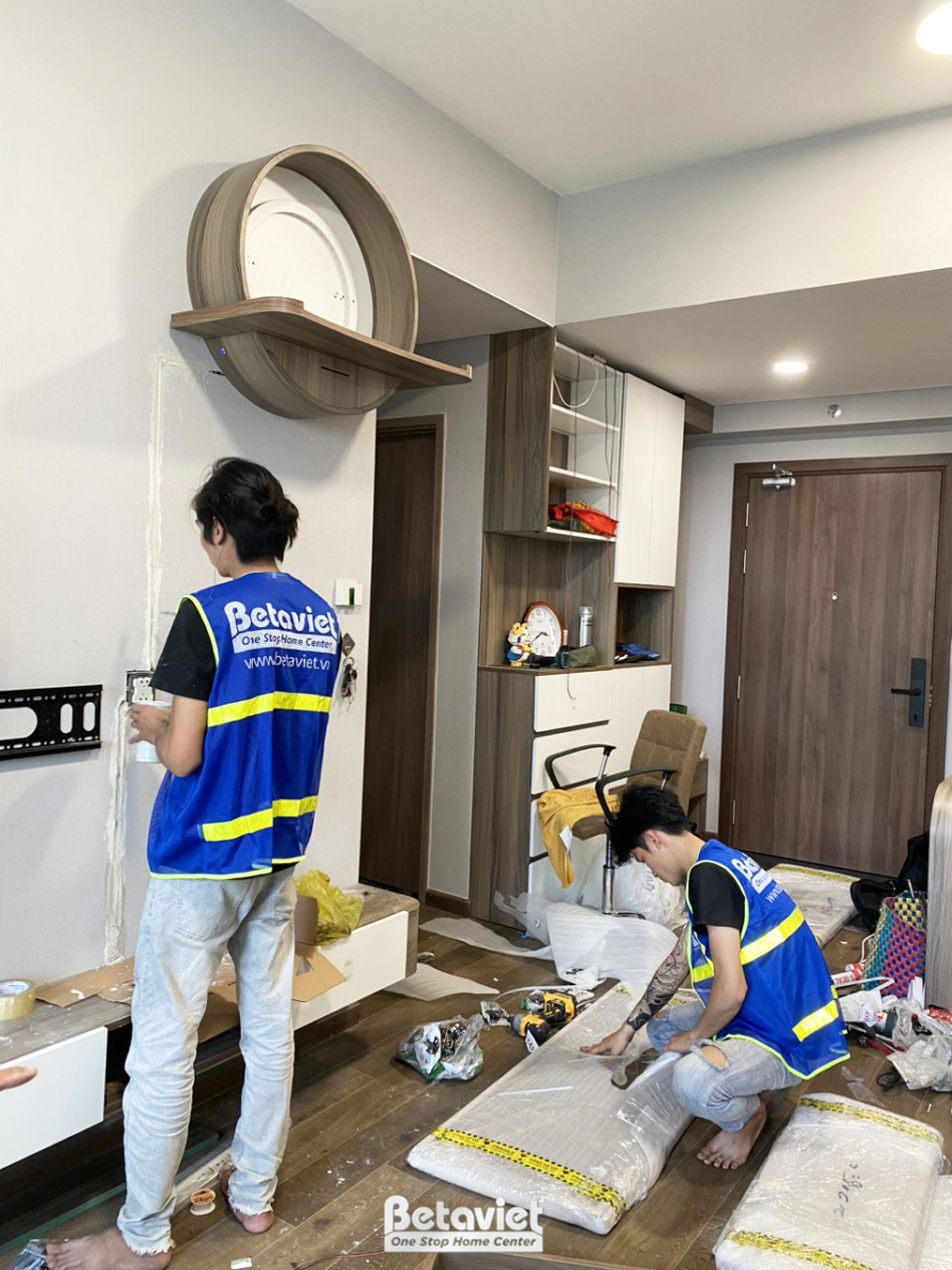 Toàn cảnh khu vực phòng khách và lối vào căn hộ đang trong quá trình lắp đặt. Các tủ kệ âm tường màu trắng và nâu gỗ được…