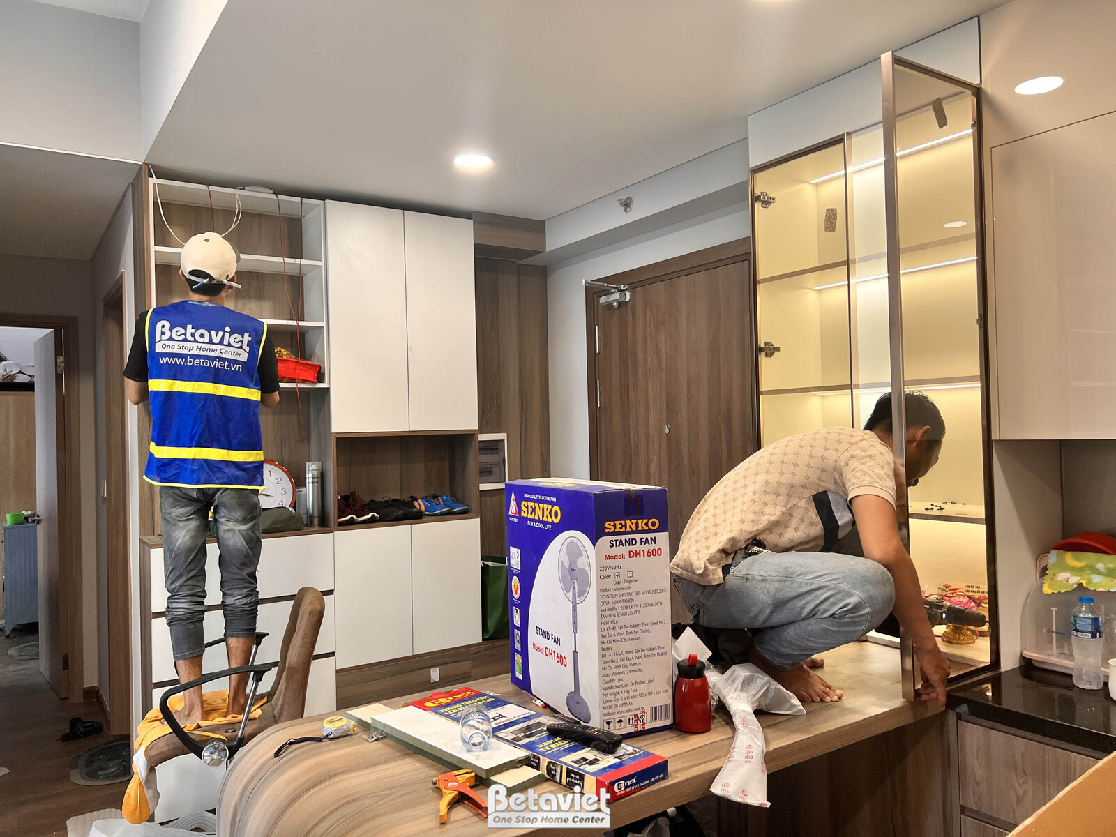 Cận cảnh khu vực tủ trang trí và tủ rượu đang được lắp đặt. Tủ gỗ công nghiệp màu trắng và nâu kết hợp các ngăn kính có đè…