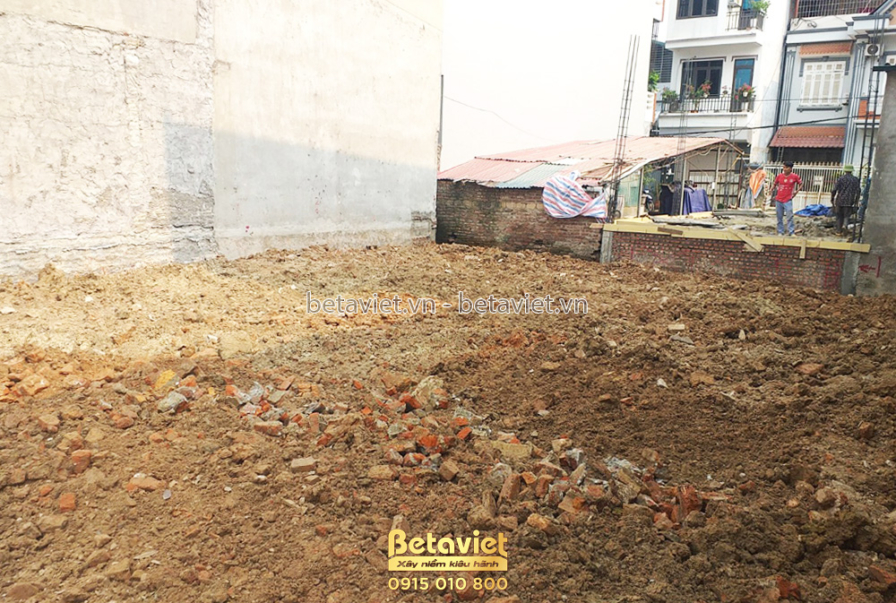 Nền đất trống đã được san phẳng, chuẩn bị cho công tác đào móng. Xung quanh là tường rào tạm và các công trình cũ. Ánh sán…