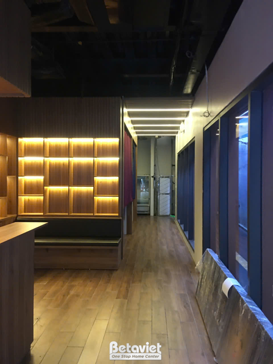Khu vực bàn ăn hoặc chờ với ghế sofa bọc da màu nâu đậm, bàn gỗ đơn giản. Tường phía sau được ốp gỗ sọc ngang và hắt sáng…