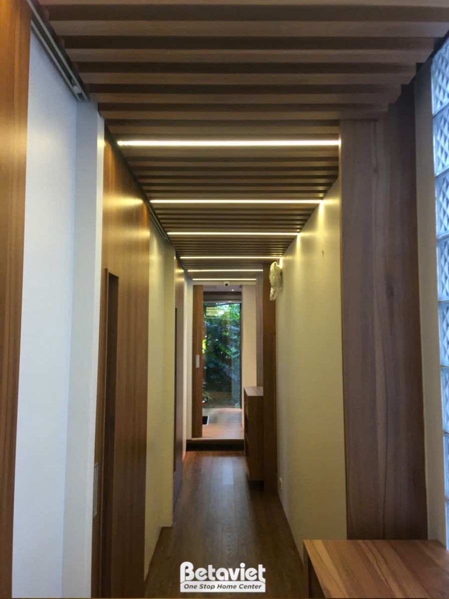 Hành lang dài với trần ốp gỗ và hệ đèn LED âm chạy dọc, tạo hiệu ứng ánh sáng dẫn lối. Tường màu be nhạt kết hợp sàn gỗ, m…