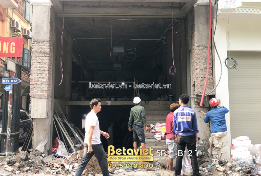 Mặt tiền nhà phố đang trong giai đoạn hoàn thiện, với cửa cuốn kim loại màu xám đóng kín. Công nhân đang làm việc ở khu vự…